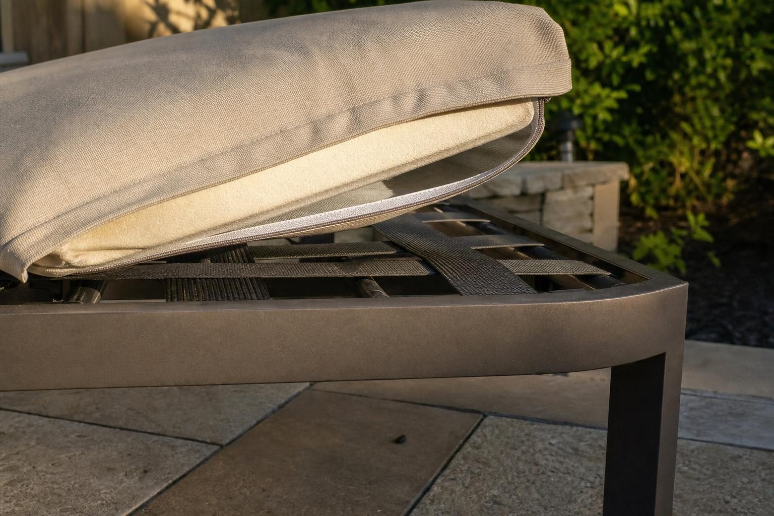 Close detail of an outdoor ottoman's frame and cushion construction, cushion partially lifted to show the support structure