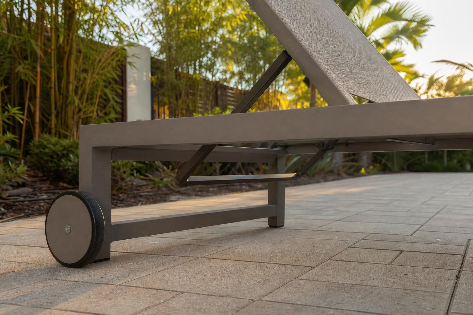Close low-angle detail of an aluminum-frame chaise lounge base with a metal wheel on one end, resting on a Florida pool deck paver surface